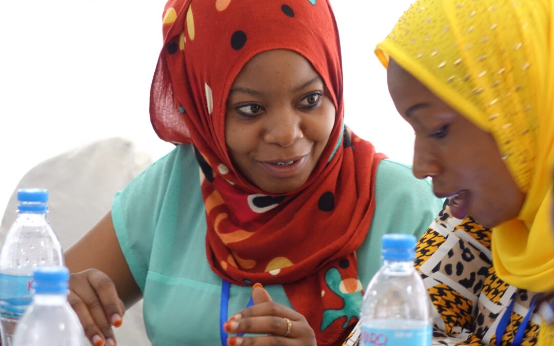 Two field workers in bright headscarves collaborate during a mobile data collection app strategy and planning, reviewing forms and discussing data entry best practices. Bottles of water are placed on the table in front of them.