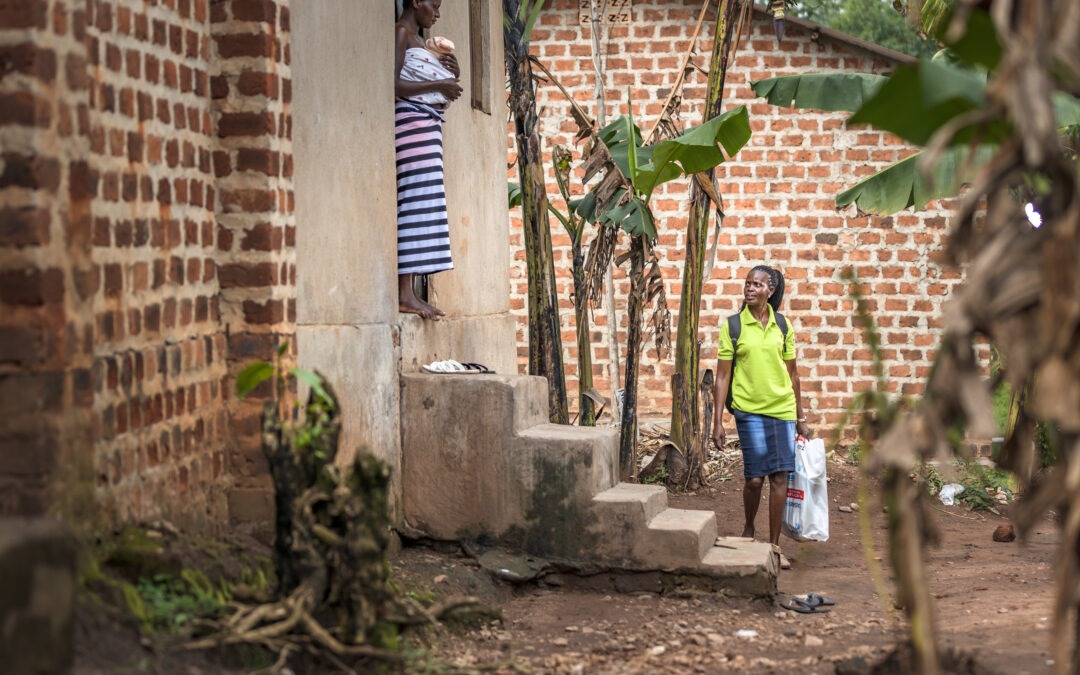 A Connect KMC frontline worker in a green shirt arrives at a rural home to provide follow-up support to a mother standing in the doorway practicing Kangaroo Mother Care with her newborn.