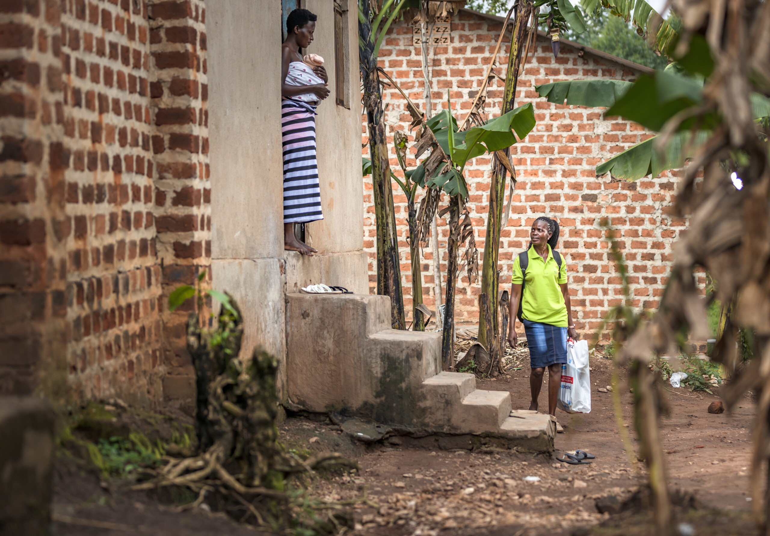 A Connect KMC frontline worker in a green shirt arrives at a rural home to provide follow-up support to a mother standing in the doorway practicing Kangaroo Mother Care with her newborn.