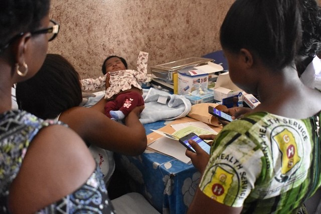 Two health workers using smartphones to record data while a mother holds her infant for an examination in a community clinic setting. One health worker wears a uniform with the Ghana Health Service.